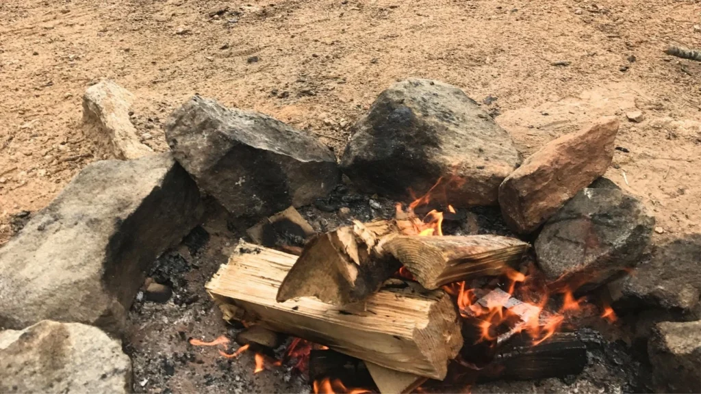 Close-up photo of logs burning in a campfire, showing glowing embers and flames at a campsite.