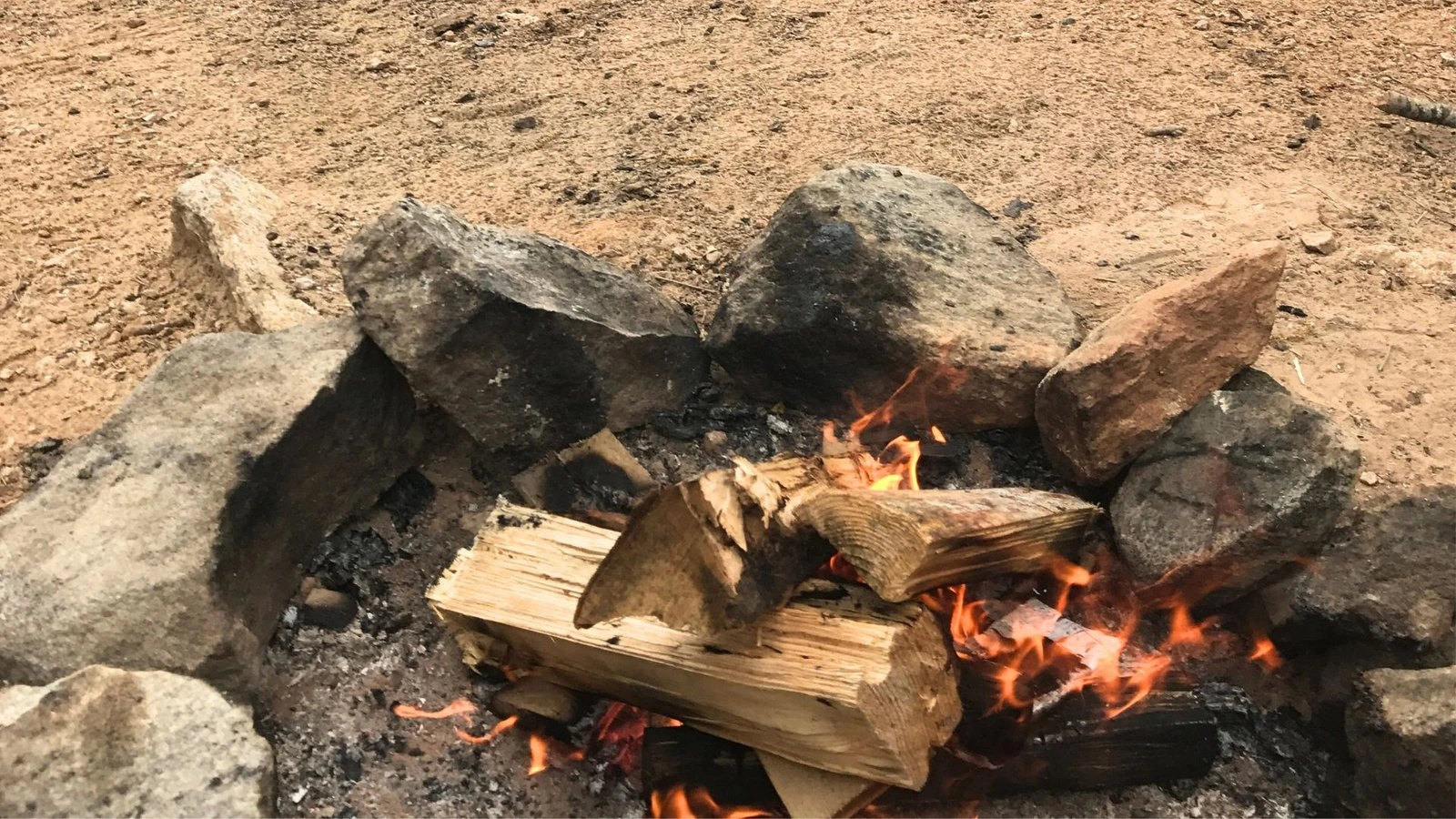 Close-up photo of logs burning in a campfire, showing glowing embers and flames at a campsite.