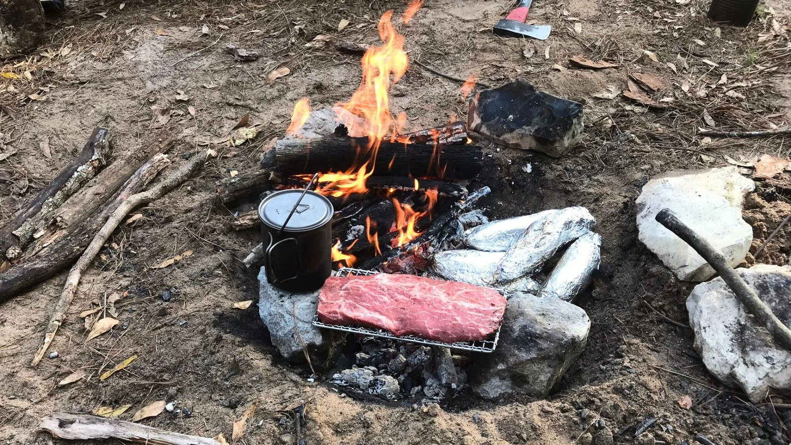 Photo of a steak cooking over a campfire grill with foil-wrapped potatoes roasting in the coals, showing a classic camping meal.