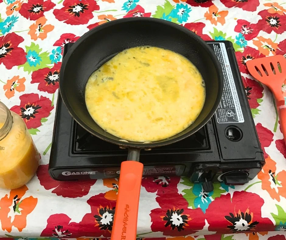 Photo of scrambled eggs cooking in a pan on a single burner camp stove, showing a simple camping breakfast setup.