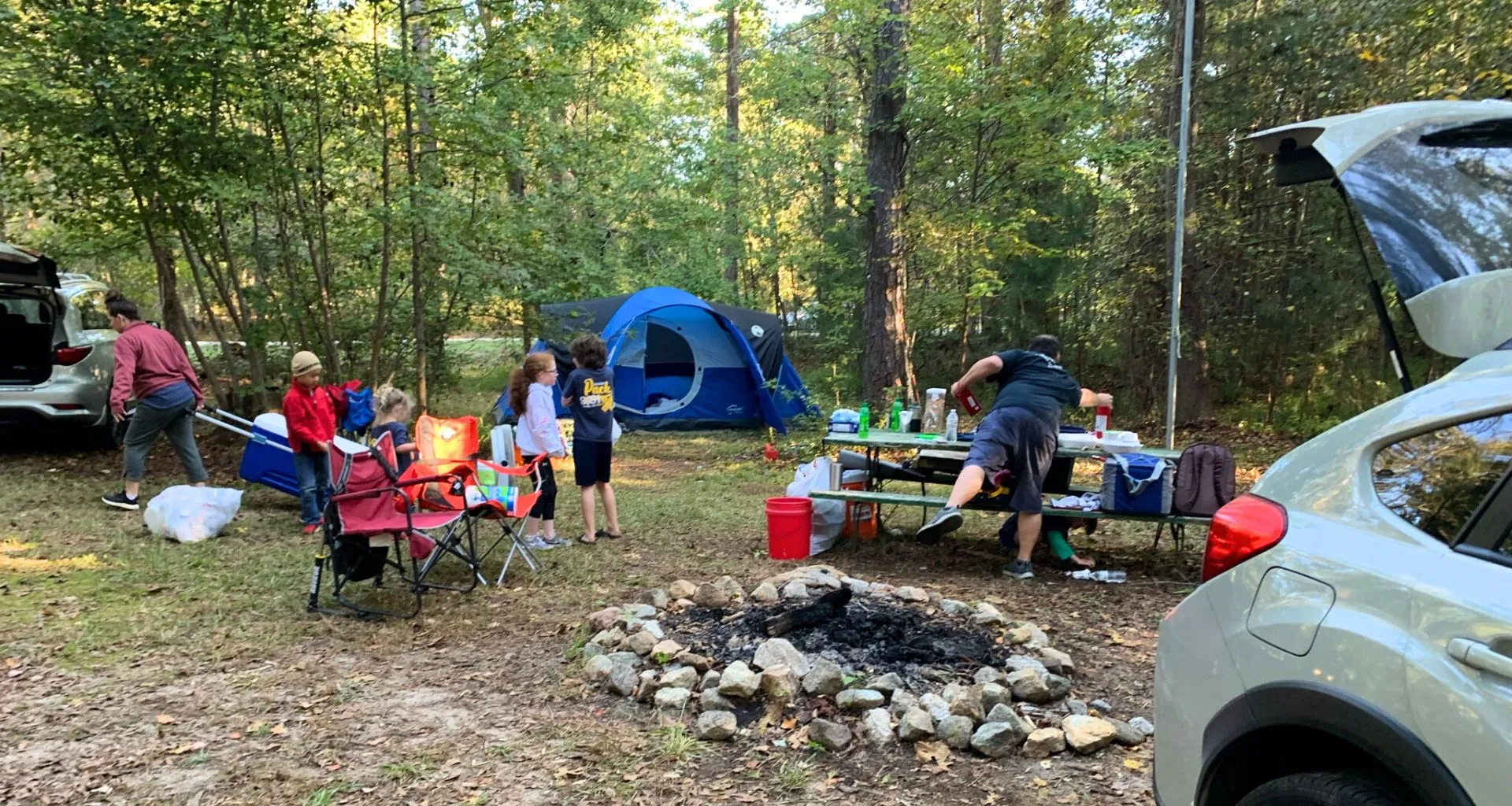 Father setting up a family campsite while several children sit in camp chairs around a fire ring